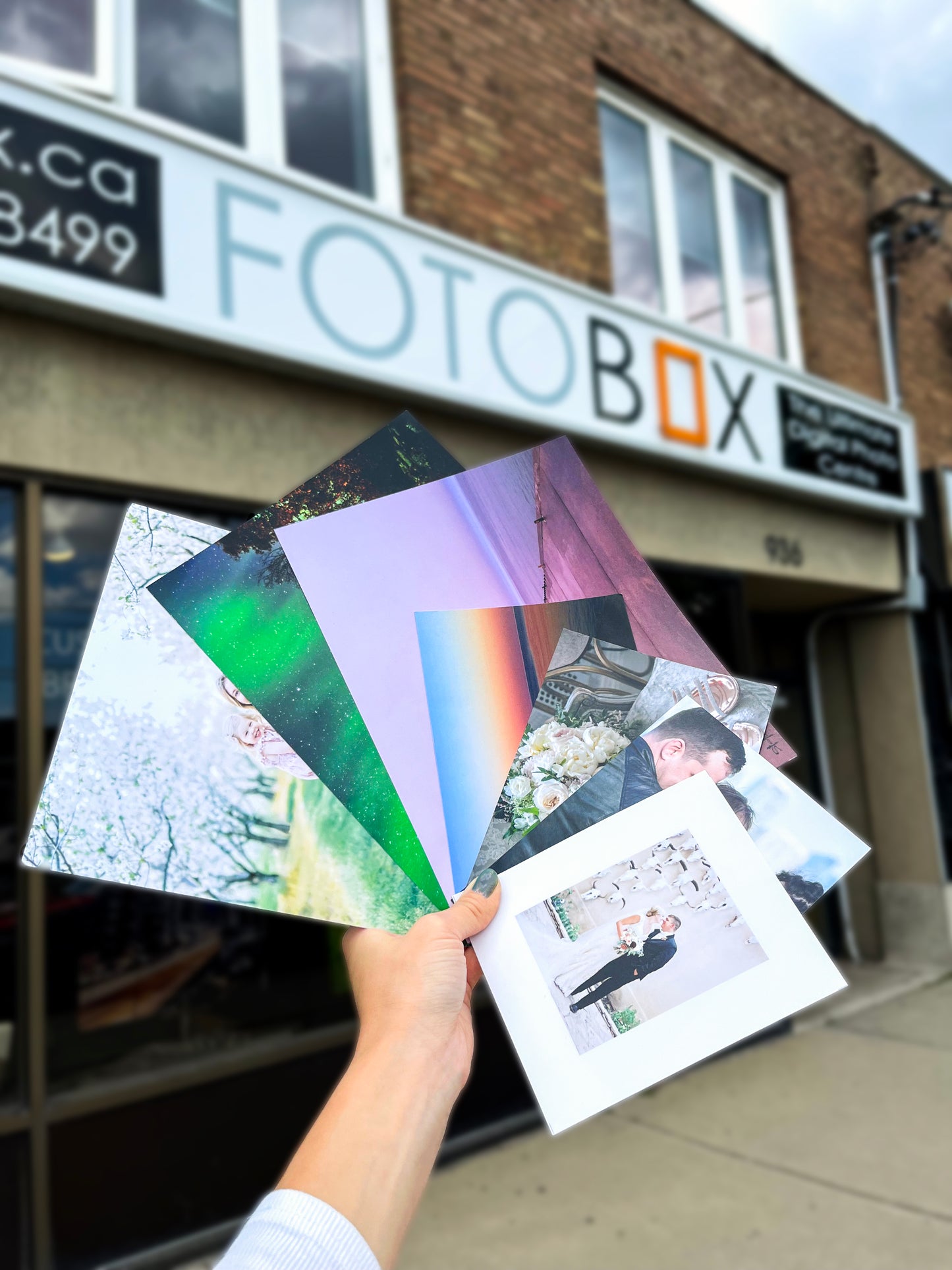 Photo prints held outside the Fotobox storefront displayed in one hand. The colors display a variety of different scenarios ranging from nature scenes, aurora borealis, family photos, and a wedding couple.