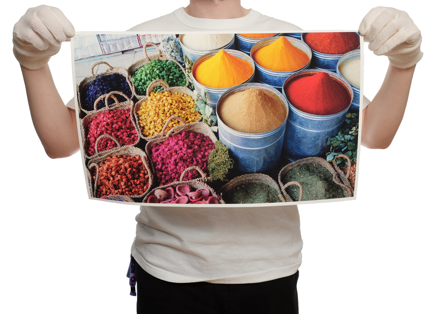 Person with white gloves and white tshirt holding up a 12x20" Giclée print. The print features a variety of vibrant and colorful yellow, deep red and beige spices filled to the brim like small hill tops in silver bins. Next to them are corded rope beige bags filled with rainbow dried herbs to display the quality of the print to the original photography.